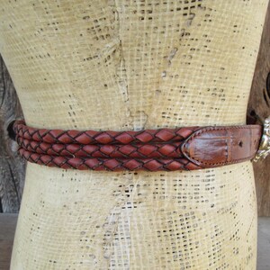 Op de afbeelding: Een bruine leren riem met een gevlochten ontwerp. De riem heeft een zilverkleurige gesp met een decoratief ontwerp. De riem wordt op een mannequin getoond.