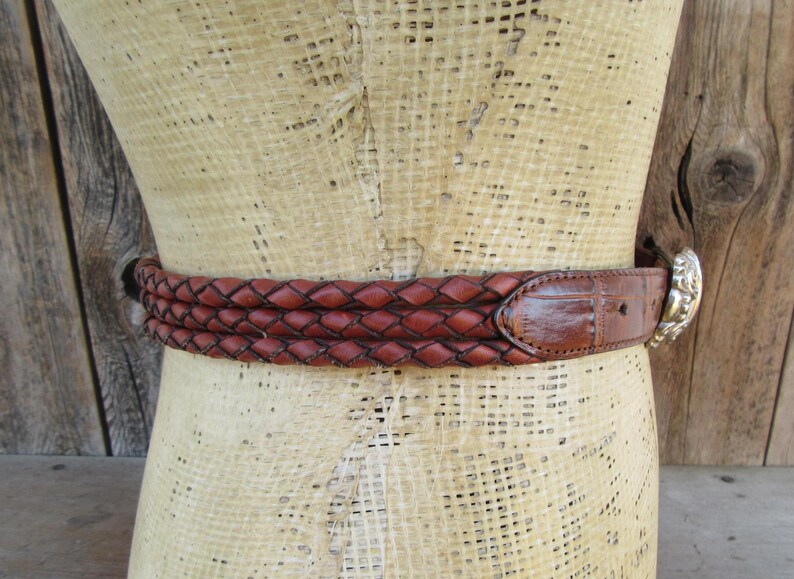 Op de afbeelding: Een bruine leren riem met een gevlochten ontwerp. De riem heeft een zilverkleurige gesp met een decoratief ontwerp. De riem wordt op een mannequin getoond.