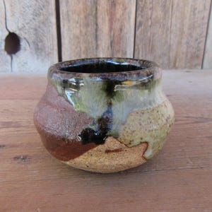 May include: A small, round, hand-thrown ceramic bowl with a wide opening. The exterior features a textured, earthy brown base that transitions into a glossy, dripped glaze in shades of dark green, cream, and tan around the upper rim.