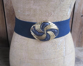 May include: A navy blue elastic belt with a large, gold-toned buckle. The buckle features a swirling, enamel design in navy and gold. The belt is displayed on a mannequin against a wooden background.