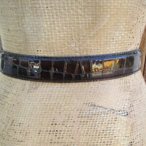 Op de afbeelding: Een donkerbruine, glanzende riem met een krokodillenleerpatroon is om een ​​mannequin gewikkeld. De riem is van gemiddelde breedte en lijkt van leer te zijn. De mannequin is lichtbruin van kleur.