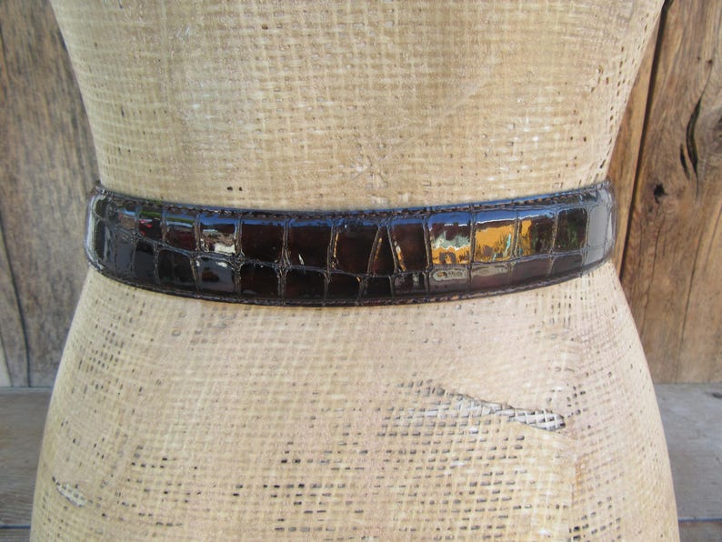 Op de afbeelding: Een donkerbruine, glanzende riem met een krokodillenleerpatroon is om een ​​mannequin gewikkeld. De riem is van gemiddelde breedte en lijkt van leer te zijn. De mannequin is lichtbruin van kleur.