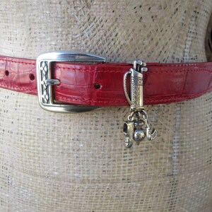 Puede incluir: Cintur&oacute;n de cuero rojo con hebilla plateada y un charm con tem&aacute;tica de golf. El cintur&oacute;n tiene un patr&oacute;n texturizado, similar al de cocodrilo. El charm plateado presenta una bolsa de golf, palos y otros accesorios de golf.