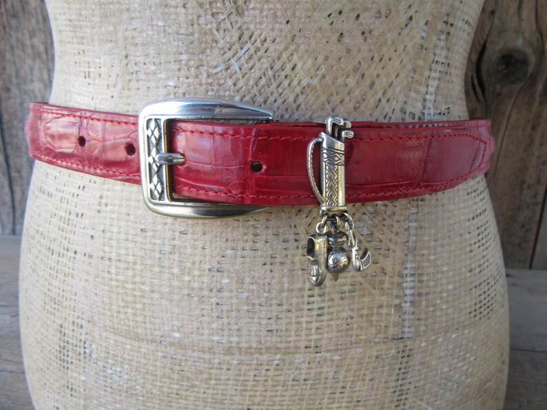 Puede incluir: Cintur&oacute;n de cuero rojo con hebilla plateada y un charm con tem&aacute;tica de golf. El cintur&oacute;n tiene un patr&oacute;n texturizado, similar al de cocodrilo. El charm plateado presenta una bolsa de golf, palos y otros accesorios de golf.