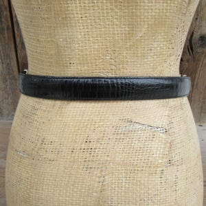 May include: A black belt with a textured, reptile-like pattern is wrapped around a mannequin. The belt is made of a shiny material and has a simple silver buckle. The mannequin is covered in a beige fabric.