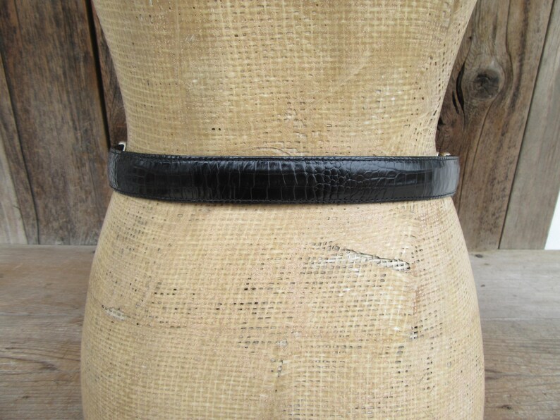 May include: A black belt with a textured, reptile-like pattern is wrapped around a mannequin. The belt is made of a shiny material and has a simple silver buckle. The mannequin is covered in a beige fabric.