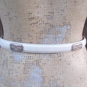 May include: A cream-colored belt with rectangular silver-toned buckles. The belt is made of a smooth material and is displayed on a mannequin. The buckles have an ornate design.