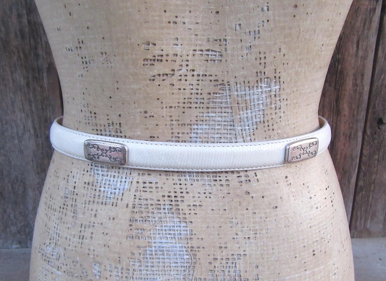 May include: A cream-colored belt with rectangular silver-toned buckles. The belt is made of a smooth material and is displayed on a mannequin. The buckles have an ornate design.