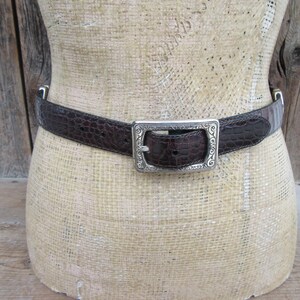 May include: A brown leather belt with a crocodile texture is displayed on a mannequin. The belt has a silver-colored rectangular buckle with an ornate design. The belt is a classic accessory.