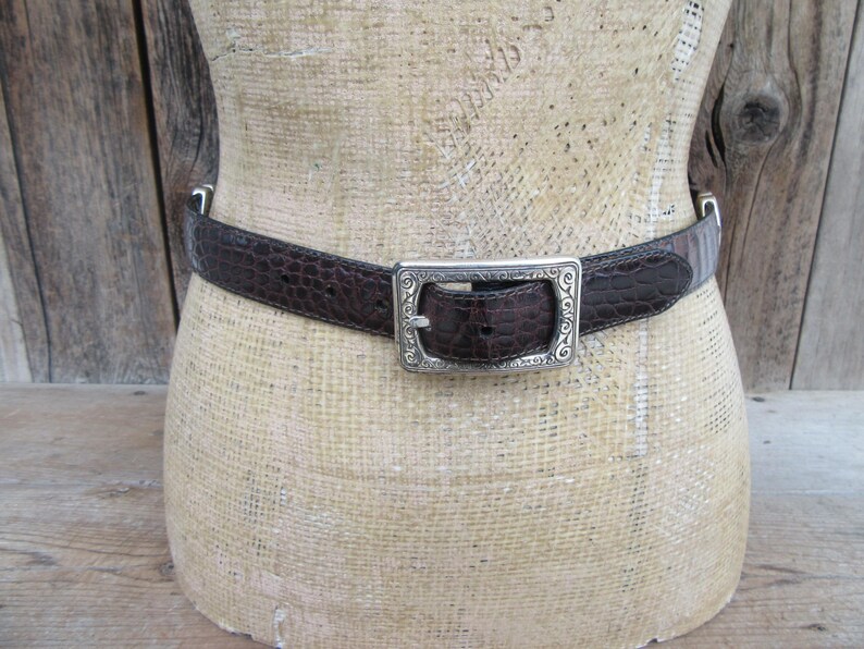 May include: A brown leather belt with a crocodile texture is displayed on a mannequin. The belt has a silver-colored rectangular buckle with an ornate design. The belt is a classic accessory.
