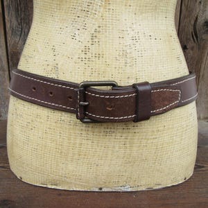 May include: A brown leather belt with white stitching and a metal buckle is displayed on a mannequin. The belt has a classic design, with a rectangular buckle and a loop to secure the belt end. The belt is approximately 1.5 inches wide.