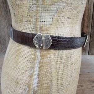 May include: A brown belt with a textured, crocodile-like pattern. The belt features a silver-colored buckle with an ornate design. The belt is displayed on a mannequin.