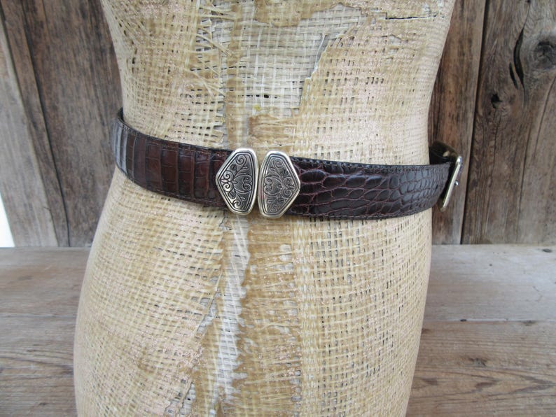 May include: A brown belt with a textured, crocodile-like pattern. The belt features a silver-colored buckle with an ornate design. The belt is displayed on a mannequin.