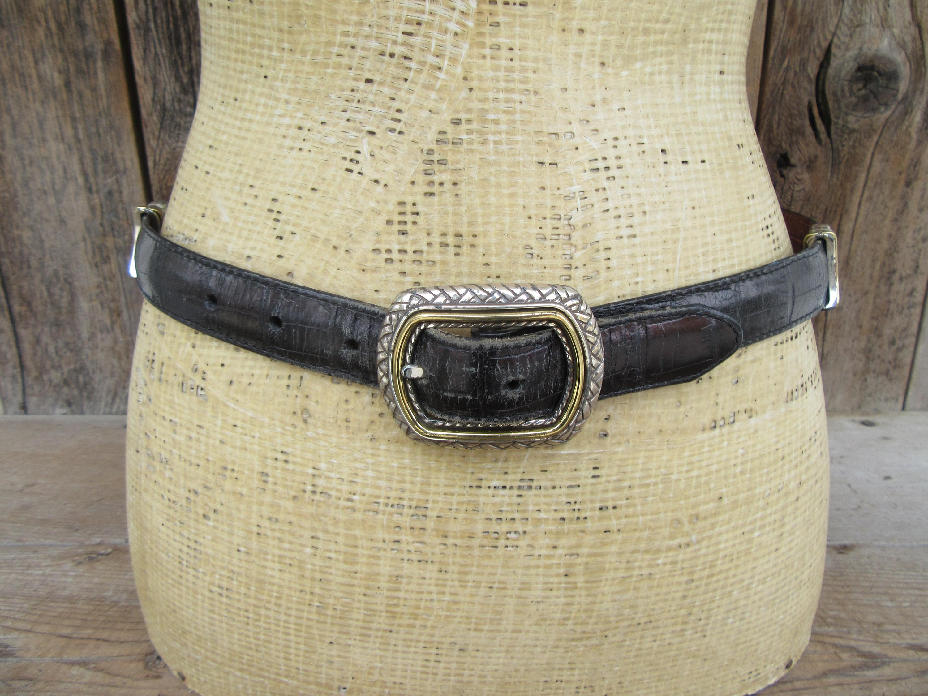 K&ouml;nnte beinhalten: Schwarzer Lederg&uuml;rtel mit silberner und goldfarbener Schnalle. Der G&uuml;rtel hat ein strukturiertes Muster und ist um eine Schaufensterpuppe befestigt. Die Schnalle ist rechteckig mit einem gewebten Design.