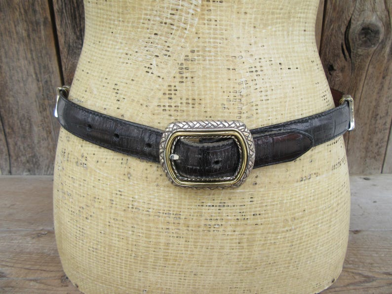 K&ouml;nnte beinhalten: Schwarzer Lederg&uuml;rtel mit silberner und goldfarbener Schnalle. Der G&uuml;rtel hat ein strukturiertes Muster und ist um eine Schaufensterpuppe befestigt. Die Schnalle ist rechteckig mit einem gewebten Design.