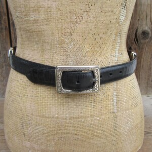 May include: Black belt with a silver buckle. The belt is made of a textured material, possibly leather, and features a rectangular silver buckle with an ornate design. The belt is displayed on a mannequin.
