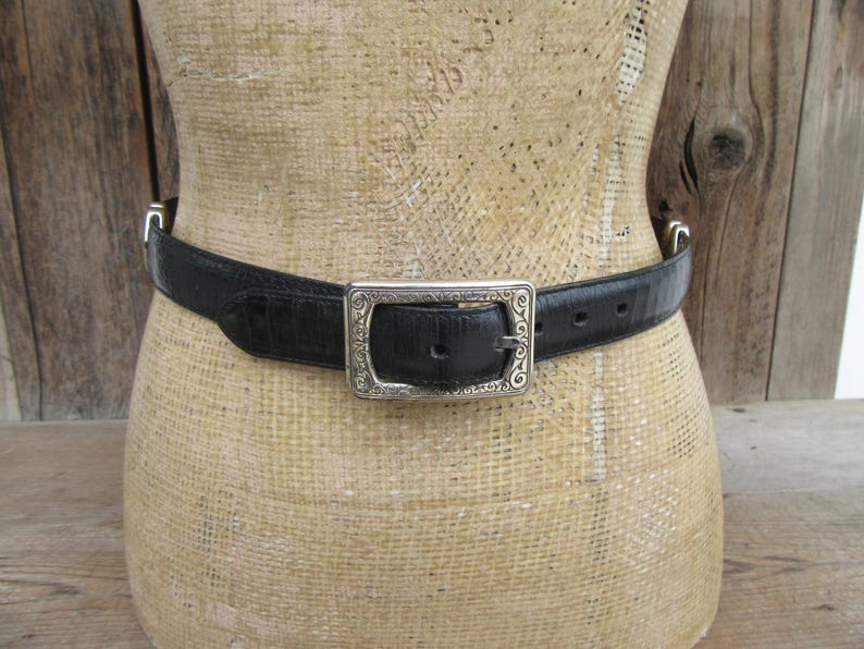 May include: Black belt with a silver buckle. The belt is made of a textured material, possibly leather, and features a rectangular silver buckle with an ornate design. The belt is displayed on a mannequin.