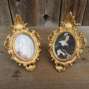 May include: Two ornate, gold-colored oval picture frames. Each frame features a portrait of a person in historical attire. The frames have a decorative, scroll-like design around the oval picture area and a decorative top.