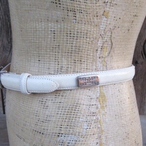 May include: A cream-colored belt with a textured surface and a silver buckle. The belt features a decorative rectangular buckle with an ornate design. The belt is displayed on a mannequin.