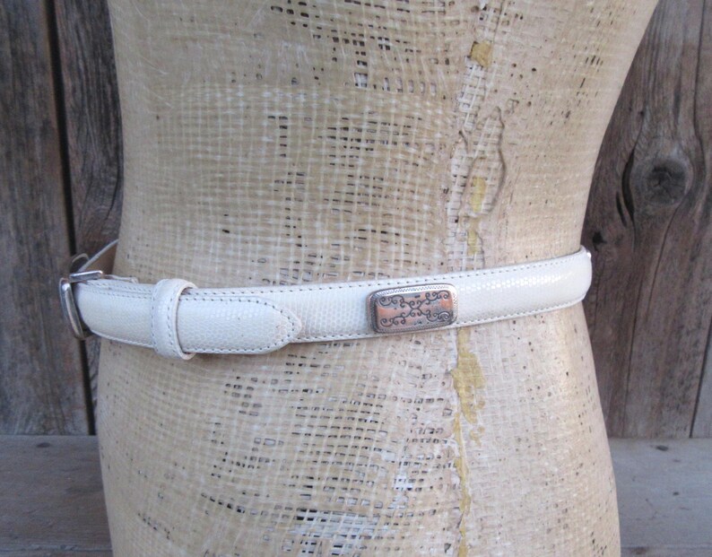 May include: A cream-colored belt with a textured surface and a silver buckle. The belt features a decorative rectangular buckle with an ornate design. The belt is displayed on a mannequin.