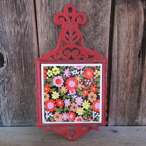 May include: A red cast iron trivet featuring an ornate, scrollwork handle and frame surrounding a square ceramic tile. The tile displays a dense, colourful floral pattern with red, pink, yellow, and white flowers set against a black background.