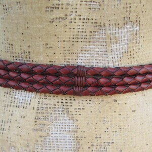 Op de afbeelding: Een gevlochten leren riem in een rijke bordeauxrode kleur. De riem bestaat uit drie verweven strengen, waardoor een textuur ontstaat. De riem wordt getoond op een mannequin, die het ontwerp en de stijl laat zien.