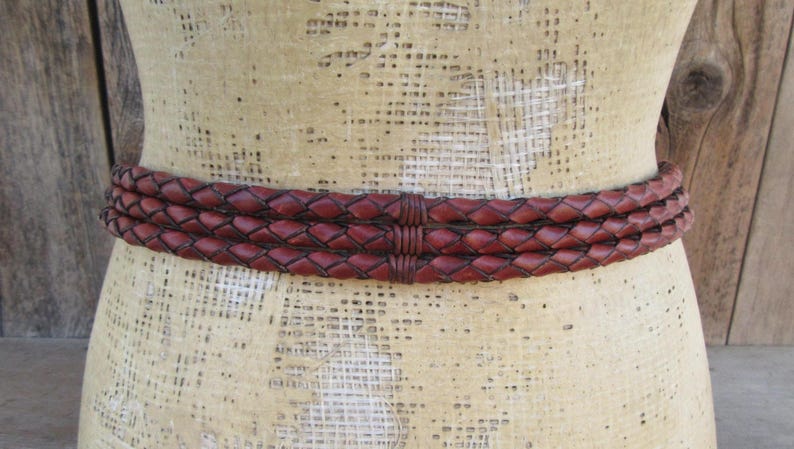 Op de afbeelding: Een gevlochten leren riem in een rijke bordeauxrode kleur. De riem bestaat uit drie verweven strengen, waardoor een textuur ontstaat. De riem wordt getoond op een mannequin, die het ontwerp en de stijl laat zien.