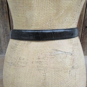May include: A black belt with a textured, crocodile-like pattern is wrapped around a mannequin. The belt is made of a shiny material and appears to be leather. The mannequin is made of a woven material.