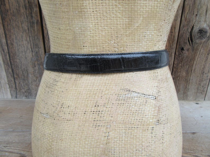 May include: A black belt with a textured, crocodile-like pattern is wrapped around a mannequin. The belt is made of a shiny material and appears to be leather. The mannequin is made of a woven material.