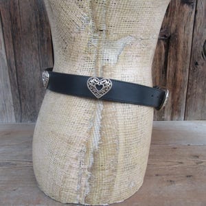 Op de afbeelding: Zwarte riem met zilveren hartvormige gesp en accenten. De riem wordt om een ​​mannequin gedragen. De hartgesp bevindt zich in het midden van de riem. De riem is gemaakt van een glad, donker materiaal.