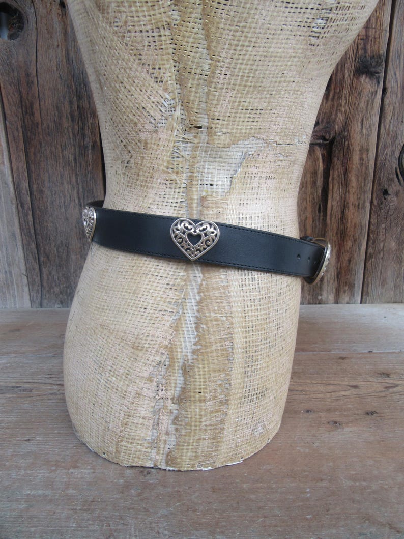 Op de afbeelding: Zwarte riem met zilveren hartvormige gesp en accenten. De riem wordt om een ​​mannequin gedragen. De hartgesp bevindt zich in het midden van de riem. De riem is gemaakt van een glad, donker materiaal.