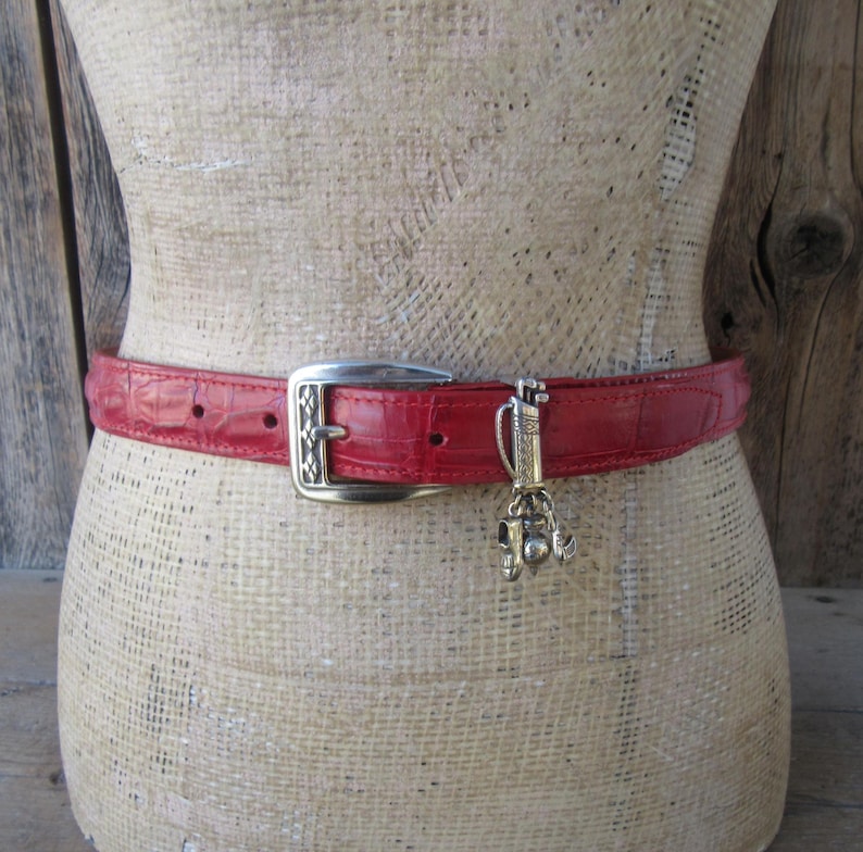 Puede incluir: Un cintur&oacute;n rojo con hebilla plateada y un charm con tem&aacute;tica de golf. El cintur&oacute;n tiene un patr&oacute;n texturizado similar al de la piel de cocodrilo. El charm presenta una bolsa de golf, palos y pelotas.