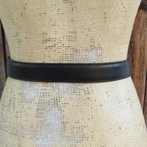 Pode incluir: Um cinto de couro preto est&aacute; enrolado em um manequim. O cinto tem um design simples e cl&aacute;ssico, com um acabamento liso e fosco. O manequim &eacute; de cor bege claro e o fundo &eacute; uma parede de madeira.