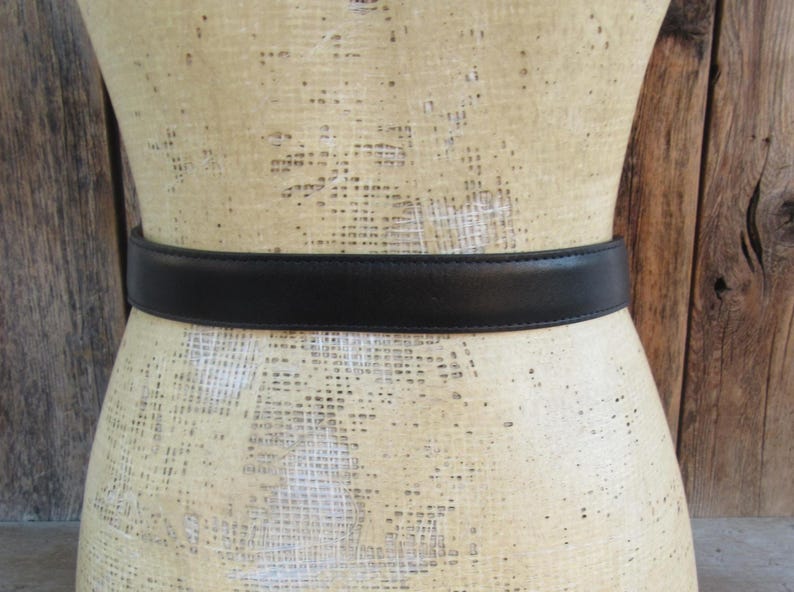 Pode incluir: Um cinto de couro preto est&aacute; enrolado em um manequim. O cinto tem um design simples e cl&aacute;ssico, com um acabamento liso e fosco. O manequim &eacute; de cor bege claro e o fundo &eacute; uma parede de madeira.