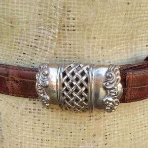 Op de afbeelding: Bruine leren riem met een zilverkleurige gesp. De riem heeft een krokodillenstructuur en een decoratieve zilveren gesp met een geweven middenontwerp. De riem is om een ​​mannequin bevestigd.