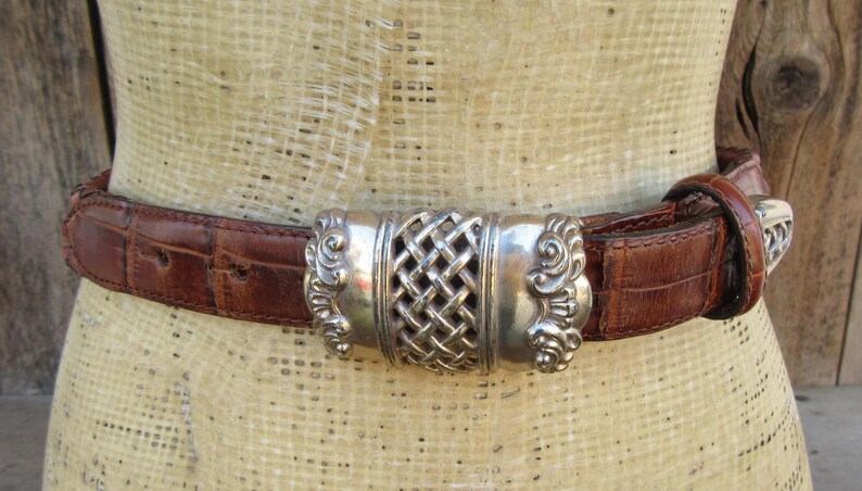 Op de afbeelding: Bruine leren riem met een zilverkleurige gesp. De riem heeft een krokodillenstructuur en een decoratieve zilveren gesp met een geweven middenontwerp. De riem is om een ​​mannequin bevestigd.