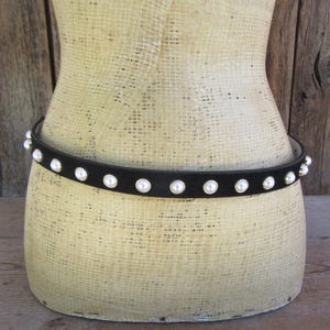 May include: A black belt with white pearl-like embellishments encircles a mannequin. The belt is made of a dark material and the pearls are evenly spaced around the circumference. The background is a rustic wooden surface.