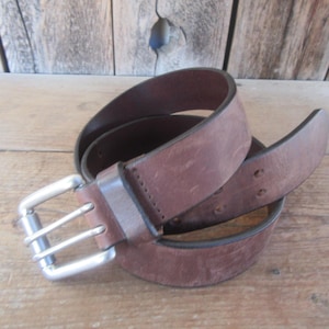 May include: A brown leather belt with a silver-colored double-bar buckle. The belt is rolled up, showing the texture and color variations of the leather. The belt is on a wooden surface.