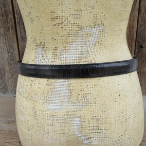 K&ouml;nnte beinhalten: Ein schwarzer G&uuml;rtel mit strukturierter Oberfl&auml;che ist um eine Schaufensterpuppe gewickelt. Der G&uuml;rtel scheint aus Leder oder einem &auml;hnlichen Material zu bestehen. Die Schaufensterpuppe ist beige und hat eine strukturierte Oberfl&auml;che.