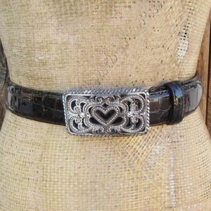 Op de afbeelding: Een donkerbruine, getextureerde leren riem met een decoratieve zilverkleurige gesp. De gesp heeft een open hartontwerp met sierlijke details. De riem wordt op een mannequin getoond.