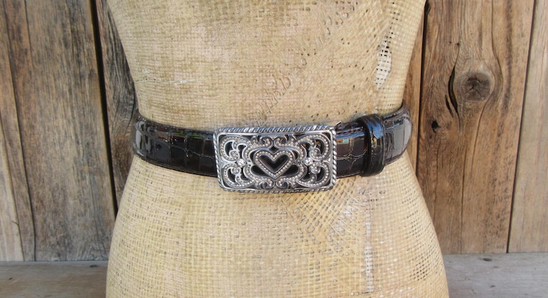 Op de afbeelding: Een donkerbruine, getextureerde leren riem met een decoratieve zilverkleurige gesp. De gesp heeft een open hartontwerp met sierlijke details. De riem wordt op een mannequin getoond.