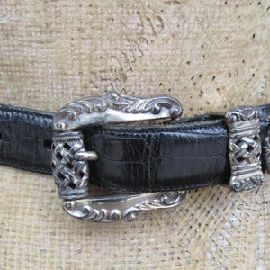 May include: A black belt with a silver buckle and accents. The belt has a textured surface and is fastened with a decorative buckle. The buckle and belt loops have an ornate design.
