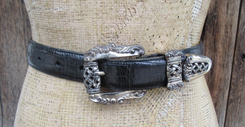 May include: A black belt with a silver buckle and accents. The belt has a textured surface and is fastened with a decorative buckle. The buckle and belt loops have an ornate design.