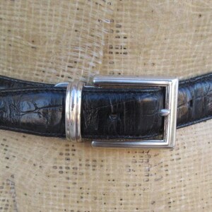 May include: Black leather belt with a silver square buckle. The belt has a textured finish and is displayed on a mannequin. The buckle is a simple, modern design. A classic accessory.