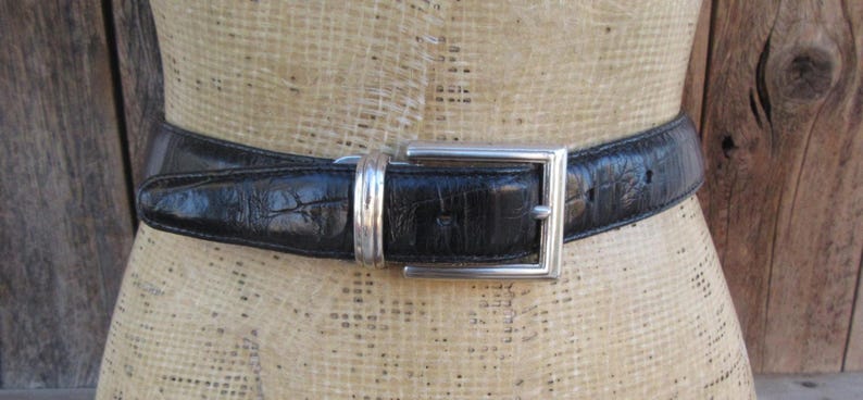 May include: Black leather belt with a silver square buckle. The belt has a textured finish and is displayed on a mannequin. The buckle is a simple, modern design. A classic accessory.