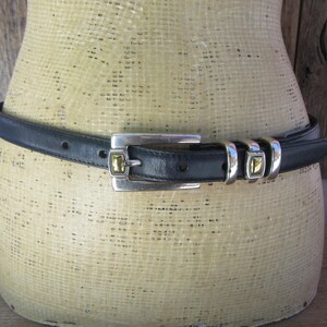 May include: A black leather belt with a silver-toned buckle and matching belt loops. The buckle features a square, gold-colored stone. The belt is displayed on a mannequin. The background is a wooden wall.