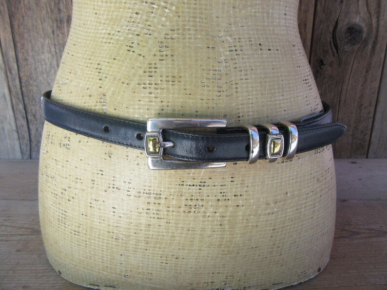 May include: A black leather belt with a silver-toned buckle and matching belt loops. The buckle features a square, gold-colored stone. The belt is displayed on a mannequin. The background is a wooden wall.