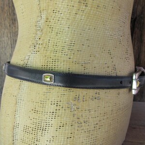 May include: Black leather belt with a silver buckle and a rectangular, light green stone set in a silver frame. The belt is displayed on a mannequin.