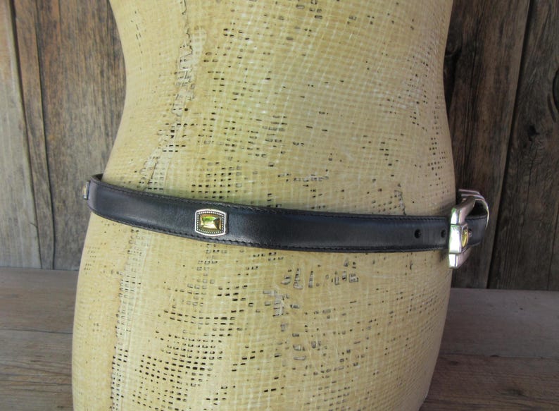 May include: Black leather belt with a silver buckle and a rectangular, light green stone set in a silver frame. The belt is displayed on a mannequin.