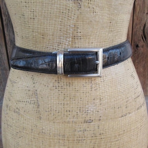 May include: A black leather belt with a silver rectangular buckle. The belt has a textured, embossed pattern. The buckle is a simple, classic design. The belt is displayed on a mannequin.